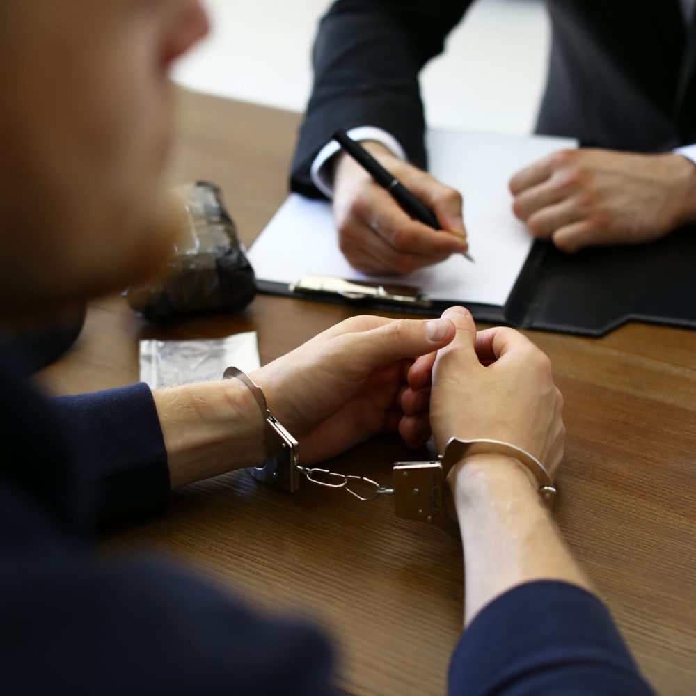 Police officer interrogating criminal in handcuffs at desk indoors