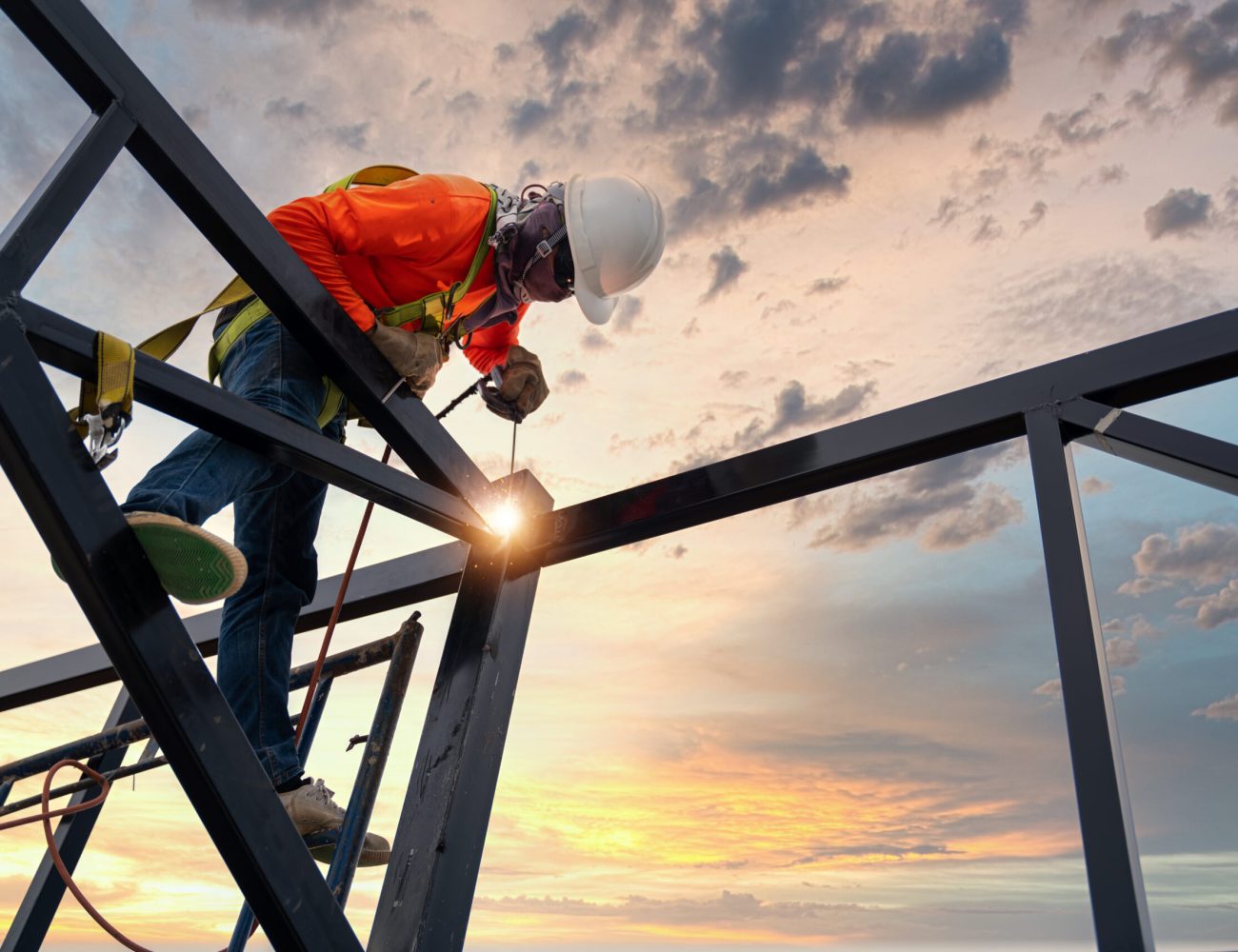 A welder is welding steel on a steel roof truss. Working at height equipment. Fall arrestor device for worker with hooks for safety body harness. Worker in construction site.